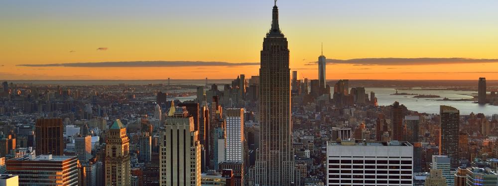 New York City skyline at sunset, featuring the Empire State Building and One World Trade Center against an orange sky.