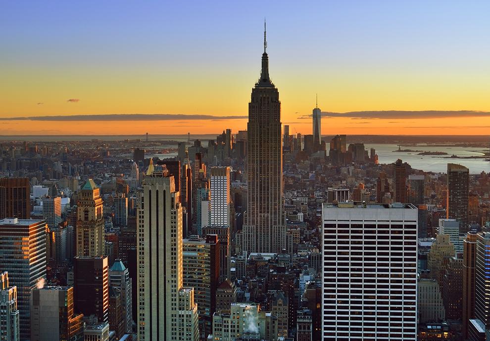 New York City skyline at sunset, featuring the Empire State Building and One World Trade Center against an orange sky.